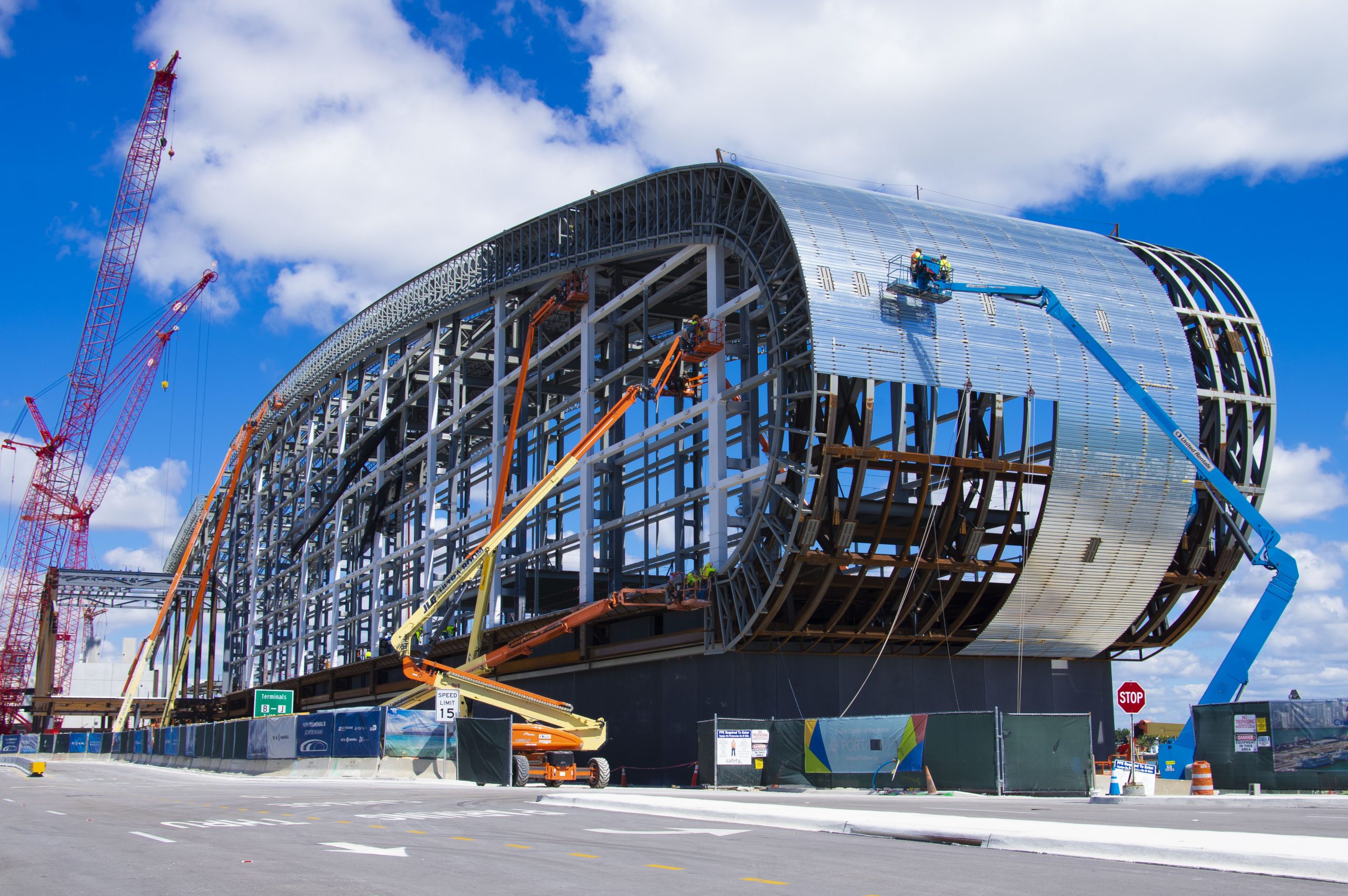 Construction of Norwegian Cruise Line Terminal B in PortMiami showing massive steel framing, cranes, and workers installing curved metal panels on the structure.